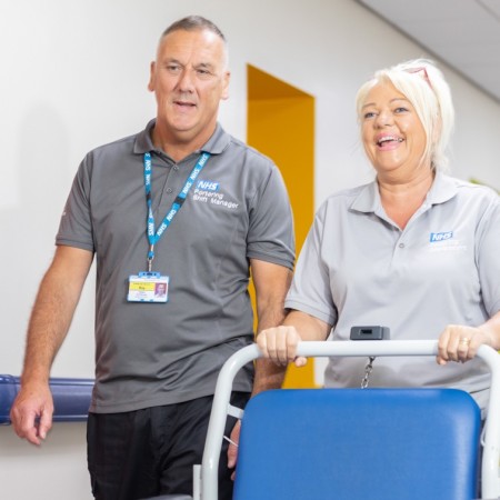 A male and female member of our Portering team walking down a hospital corridor - one pushing a patient chair.